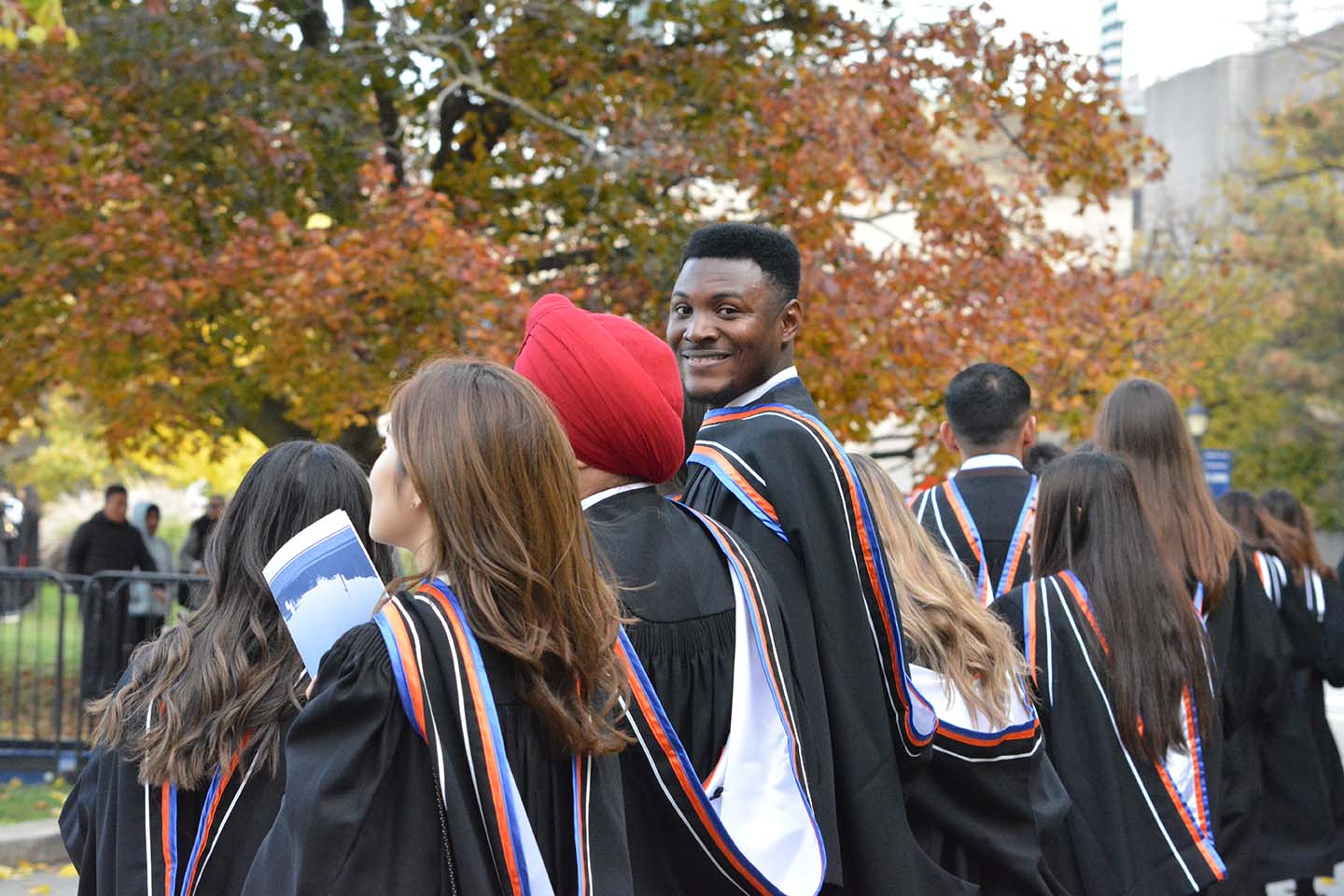 Smilling graduate standing outside amidst other graduates with orange fall trees in the background